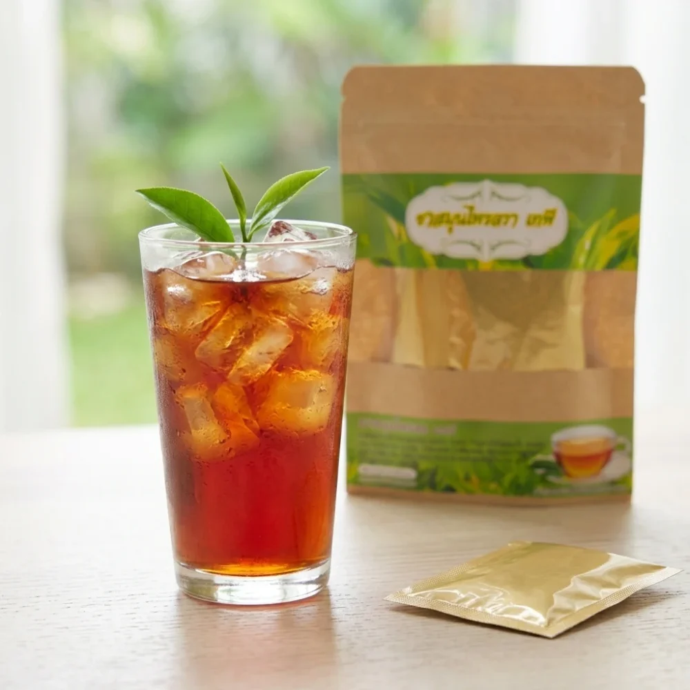15-herb Thai botanical blend tea served iced in a tall glass with ice and fresh leaves, kraft pouch and gold-foil sachet beside it
